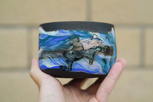 Black ceramic bowl with blue swirled coloring and a shiny abstract design of what looks like a person riding on a horse.