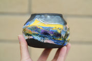 Ceramic pot with many colors, mostly blues and yellows, and shining elements, looking like an abstract desert landscape.