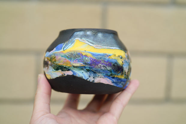 Ceramic pot with many colors, mostly blues and yellows, and shining elements, looking like an abstract desert landscape.