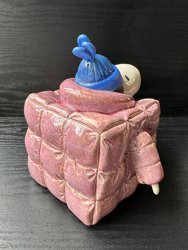 Ceramic sculpture of Snoopy wearing a puffy red jacket and blue beanie. His body is shaped like a cube.