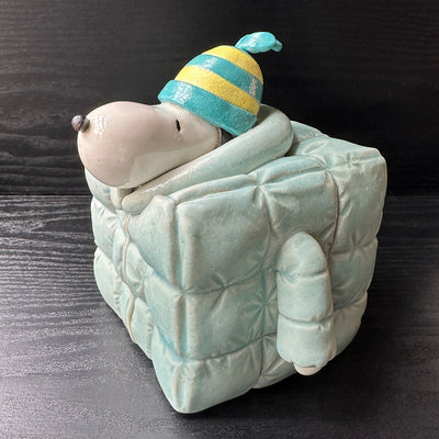 Ceramic sculpture of Snoopy wearing a puffy teal colored jacket and blue and yellow striped beanie. His body is shaped like a cube.