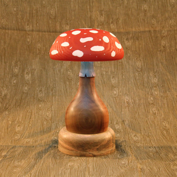 Carved wooden mushroom, with a thick and smooth wooden base. The top of the mushroom is red with while polka dots. Sculpture is also a lamp, emitting a dim glow.