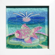 Embroidery of a pink and green goldfish inside of a flower shaped container of water. It sits atop a green lily pad with a blue gradient background. Piece is in thick white frame.