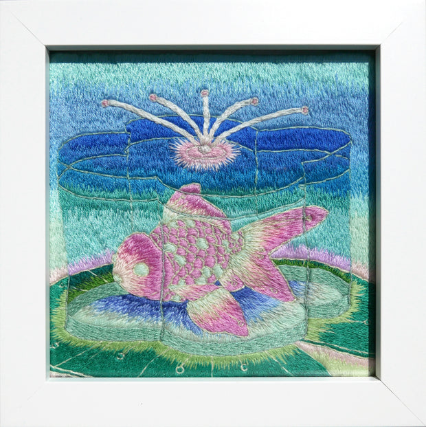 Embroidery of a pink and green goldfish inside of a flower shaped container of water. It sits atop a green lily pad with a blue gradient background. Piece is in thick white frame.