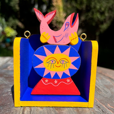 Painted wood sculpture of a sun person holding up a pink fish. Both are within an open wood box, blue with yellow trim and gold hooks to hang.