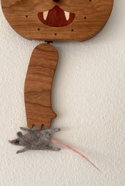 Die cut wooden sculpture, of a cat's head with a swinging wooden cat arm below it. A felt dead mouse is attached to the cats paw and the face of the cat resembles a simplified clock.