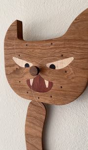 Die cut wooden sculpture, of a cat's head with a swinging wooden cat arm below it. A felt dead mouse is attached to the cats paw and the face of the cat resembles a simplified clock.