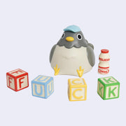 Vinyl figure of a chubby gray pigeon, sitting on the ground and wearing a small blue cap. Colorful letter blocks in front of it spell out "FUCK." A small Yakult style bottle is nearby with "Felicia Chiao" written on it.