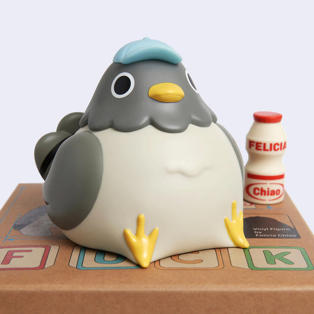 Vinyl figure of a chubby gray pigeon, sitting on its product packaging box and wearing a small blue cap. A Yakult style bottle is nearby with "Felicia Chiao" written on it.