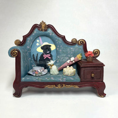Painting of a small black dog in a wide brimmed yellow hat with a pink feather coming out of it. Its framed in a 3D sculptural frame of a couch with little knick knacks on it.