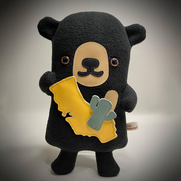 Flat plush black bear, with shiny eyes and short limbs. It holds up a plush cut out of California and a small cactus.