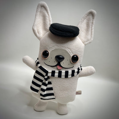 Cream colored plush dog mostly flat with large pointed ears and a sewn on face. It wears a hat and a striped scarf and has its small tongue out, smiling.