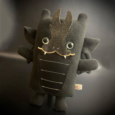 Plush of a black dragon, with a very squarish body and small arms and legs. Its facial features are made of vinyl, with a sharp tooth smile and a striped underbelly.