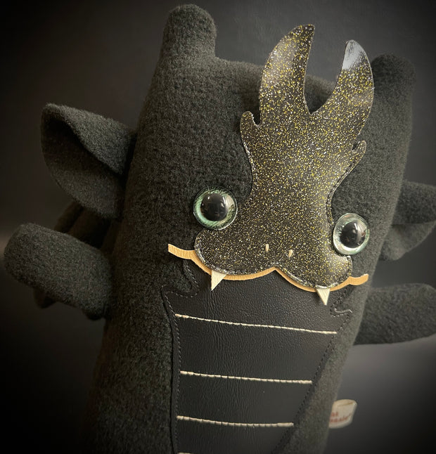 Plush of a black dragon, with a very squarish body and small arms and legs. Its facial features are made of vinyl, with a sharp tooth smile and a striped underbelly.