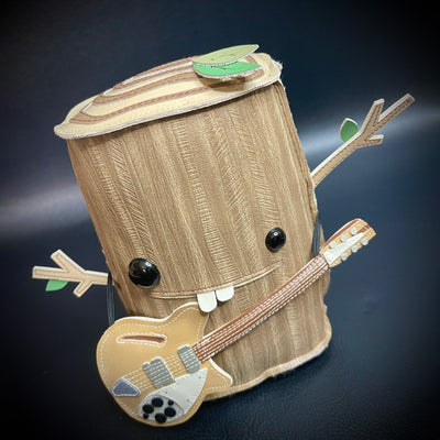 Vinyl fabric plush of a tree stump, with different sized shiny eyes and a buck tooth smile. It has its branch arms extended out in excitement and has a guitar strapped around its body.