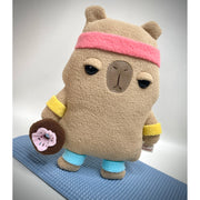 Plush of a flat shaped cartoon capybara, with a frustrated facial expression. It stands on a yoga mat with sweatbands on its head, arms and legs and holds a donut.
