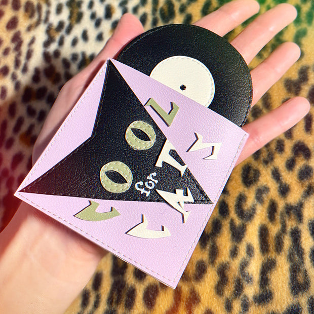 A vinyl fabric sculpture of a vinyl record, lavender purple with text that reads "Cool for Cats." Its held in someones hand.