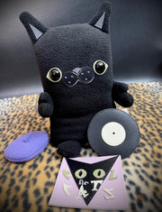 Black plush sculpture of a flat cat, with a purple beret and a vinyl sleeved record at its feet that reads "Cool for Cats." Its arms are posed in a welcoming, excited way.