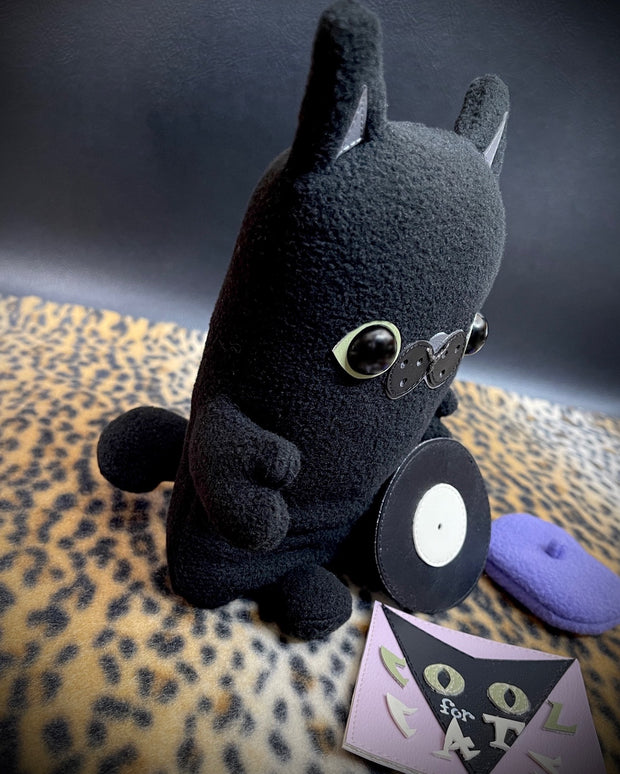 Black plush sculpture of a flat cat, with a purple beret and a vinyl sleeved record at its feet that reads "Cool for Cats." Its posed sitting on the ground holding the record.