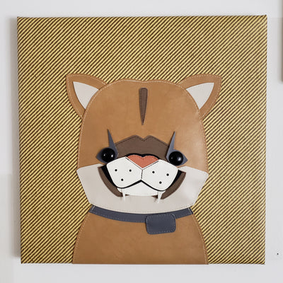 Vinyl fabric sculpture on a flat striped tan square canvas of a cartoon style baby mountain lion, with only its upper body visible.