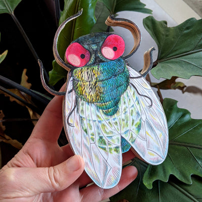 A hand holds a 6.5" fly illustration which is mounted on plywood. The fly has red eyes and a cute, pensive expression. The wings are rainbowy