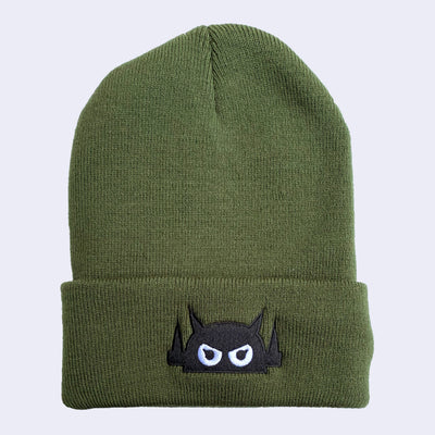 An olive green knit beanie with a robot head stitched on the cuff in black thread, with white eyes.