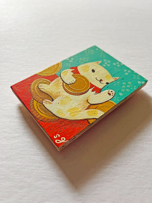     Ghahremani-yokai-maneki-neko3  576 × 576px  Painting of a chubby cream colored cat with spots holding a gold coin and standing atop of more coins. It has a red collar around its neck and is in a blue room with red floors.