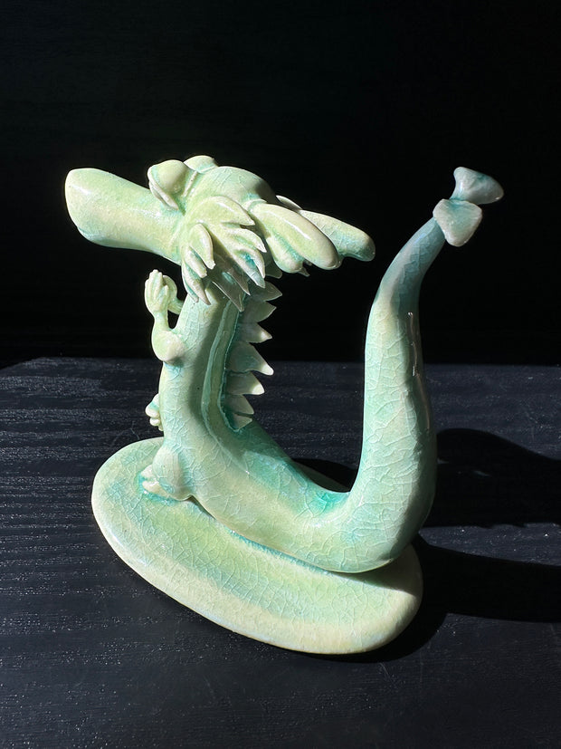 Jade colored ceramic sculpture of a dragon with a large front snout, positioned in a yoga pose with one leg up and its hands at its chest.