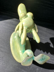 Jade colored ceramic sculpture of a dragon with a large front snout, positioned in a yoga pose with one leg up and its hands at its chest. Its tail is a sprouted leaf.