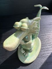 Jade colored ceramic sculpture of a dragon with a large front snout, positioned in a yoga pose with one leg up and its hands at its chest. Its tail is a sprouted leaf.
