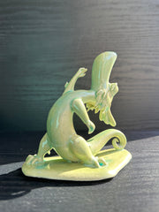 Jade colored ceramic sculpture of a dragon with a large front snout, positioned in a yoga pose with one leg in front of the other and its back bent, with one arm reaching up towards the sky.