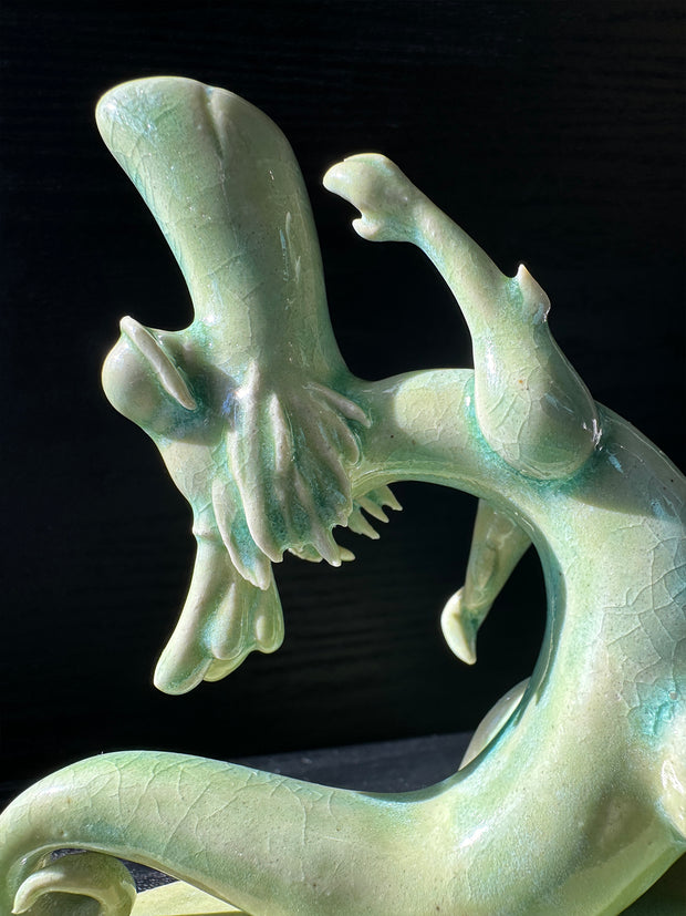 Jade colored ceramic sculpture of a dragon with a large front snout, positioned in a yoga pose with one leg in front of the other and its back bent, with one arm reaching up towards the sky.