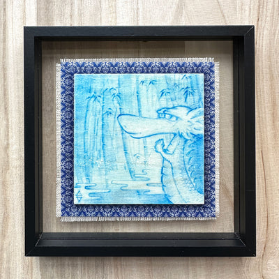 Illustration done in blue on wood board of a dragon with a long snout, holding its hands together in a bamboo forest with a pond. On a blue cloth and within a clear frame.