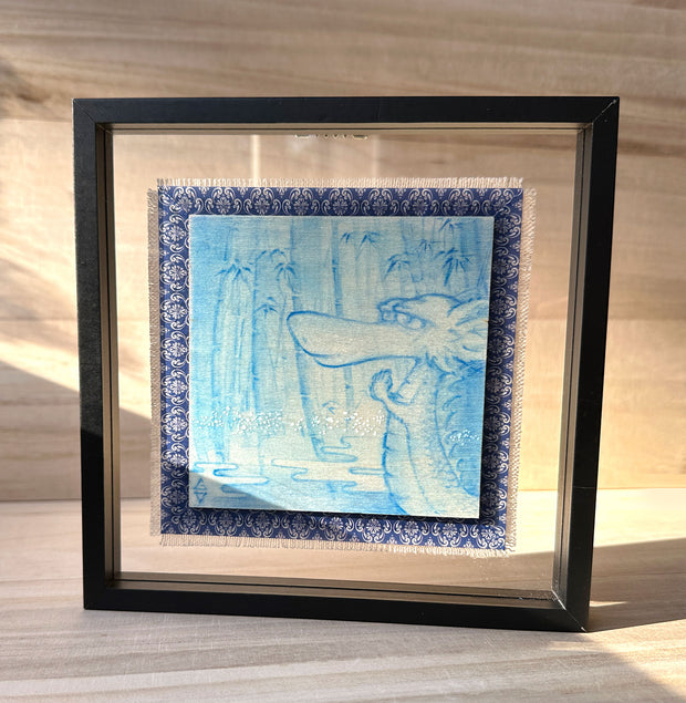 Illustration done in blue on wood board of a dragon with a long snout, holding its hands together in a bamboo forest with a pond. On a blue cloth and within a clear frame.