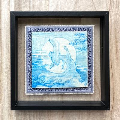 Illustration done in blue on wood board of a dragon with a long snout, doing a yoga stretch in front of a large full moon. On a blue cloth and within a clear frame.