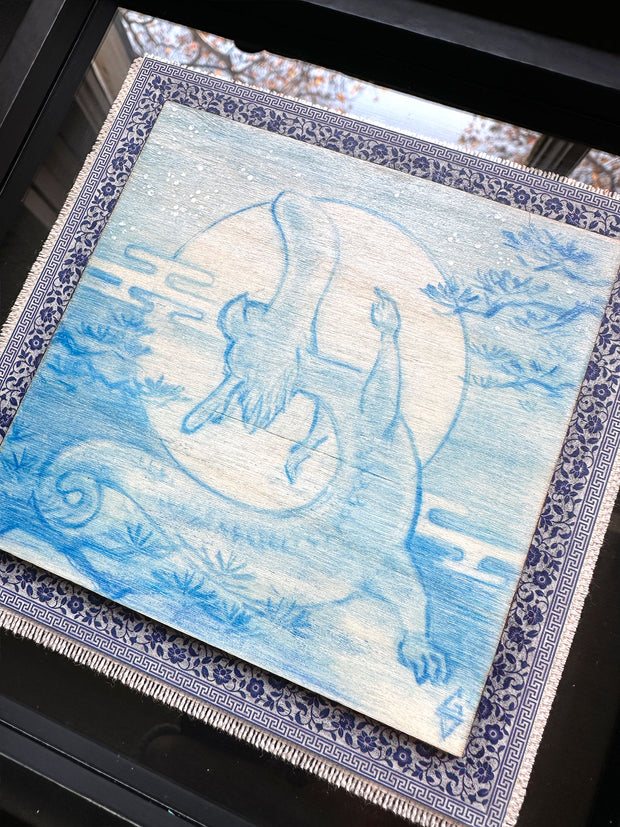 Illustration done in blue on wood board of a dragon with a long snout, doing a yoga stretch in front of a large full moon. On a blue cloth and within a clear frame.