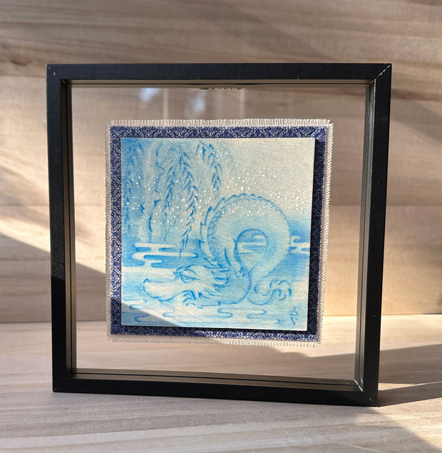 Illustration done in blue on wood board of a dragon with a long snout, doing a yoga stretch under a willow like tree. Wood board is on a blue cloth and within a clear frame.