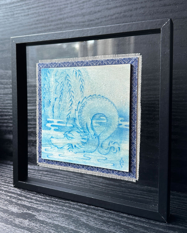 Illustration done in blue on wood board of a dragon with a long snout, doing a yoga stretch under a willow like tree. Wood board is on a blue cloth and within a clear frame.