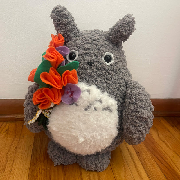 Plush doll of Totoro holding a bouquet of orange and purple flowers.