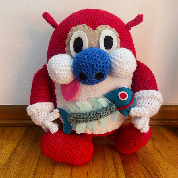Crocheted sculpture of Stimpy from Ren and Stimpy. He sits with his tongue out comically and holds a fish skeleton in his hands.