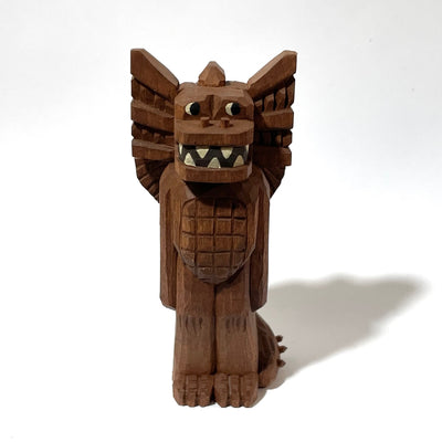Whittled wooden sculpture of a standing dragon, with large ears, a sharp tooth mouth and a long spiked tail. It looks off to the side and has its arms at its side as it stands straight.