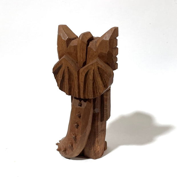 Whittled wooden sculpture of a standing dragon, with large ears, a sharp tooth mouth and a long spiked tail. It looks off to the side and has its arms at its side as it stands straight.