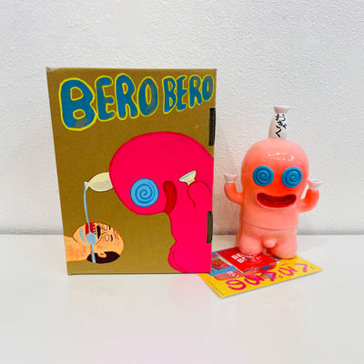 Light pink soft vinyl figure of a chubby character with a large, round head. It has a smiling face and swirled blue eyes. Atop its head is a bottle of sake, with a cup in each of its hands. It stands next to a painted box.