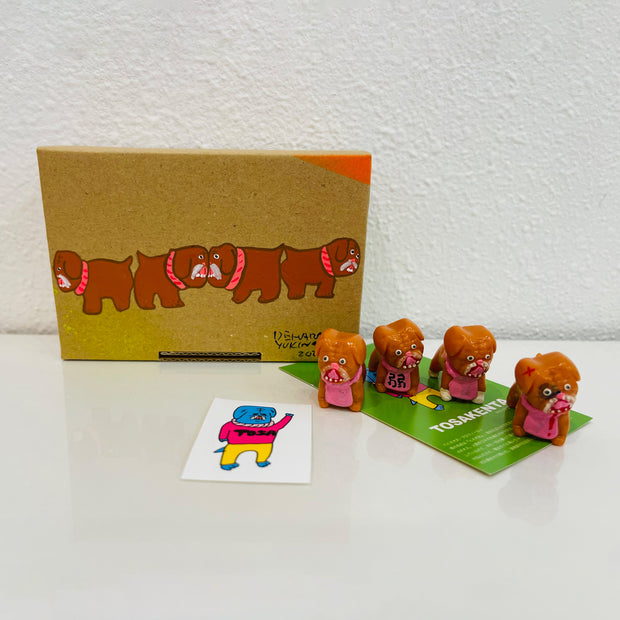4 small, brown soft vinyl bulldogs with their mouths open and tongues out. They each have bright pink eyes and pink fabric around their neck, some with writing, and some blank. They stand in front of their product packaging.