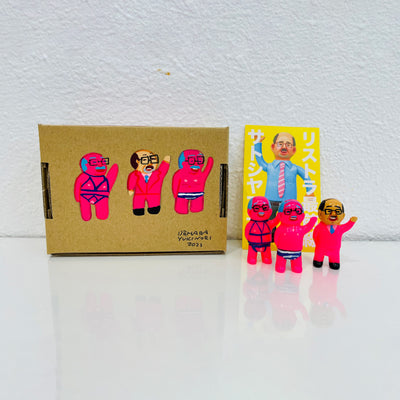 Set of 3 small vinyl neon pink figures of little business men. One wears a suit, another wears a red bondage wear and the last wears striped underwear. They stand next to a painted box.