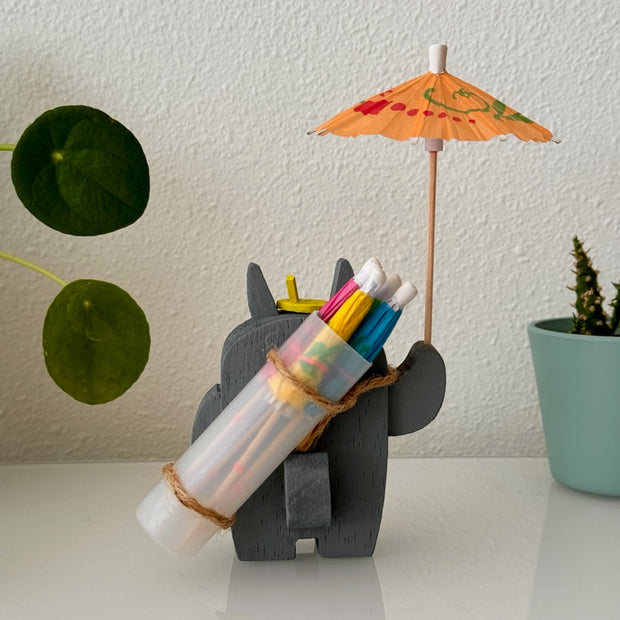  Small wooden sculpture of a smiling Totoro, looking off to the side. He holds up a small cocktail umbrella, with an over the shoulder bag holding 4 more umbrellas.