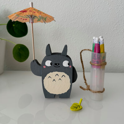 Small wooden sculpture of a smiling Totoro, looking off to the side. He holds up a small cocktail umbrella, with an over the shoulder bag holding 4 more umbrellas.