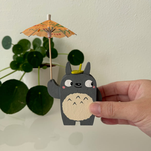  Small wooden sculpture of a smiling Totoro, looking off to the side. He holds up a small cocktail umbrella and has a wooden leaf atop his head.
