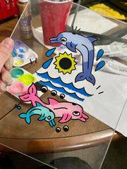  Painting on glass of 3 dolphins: a larger blue one, swimming over waves and 2 more smaller dolphins, one pink and one teal blue. A sun akin to a sunflower hangs in the sky below the first jumping dolphin.