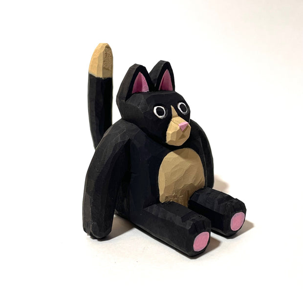 Whittled wooden sculpture of a fat black cat with pink ears and bottom of feet. It has a tan snout, belly and tail and looks straight at the viewer with its mouth slightly ajar, surprised looking.